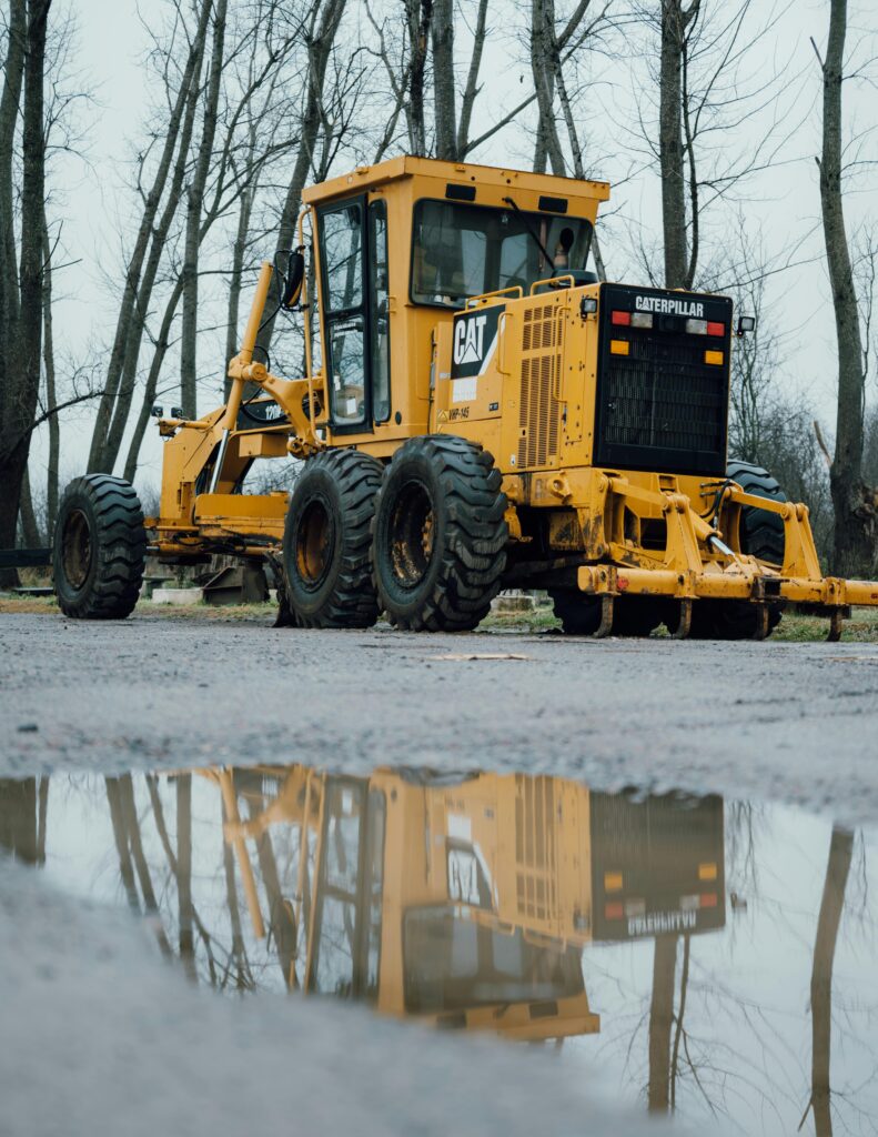 Yellow grader parked on a construction site with reflection in a puddle, surrounded by bare trees.